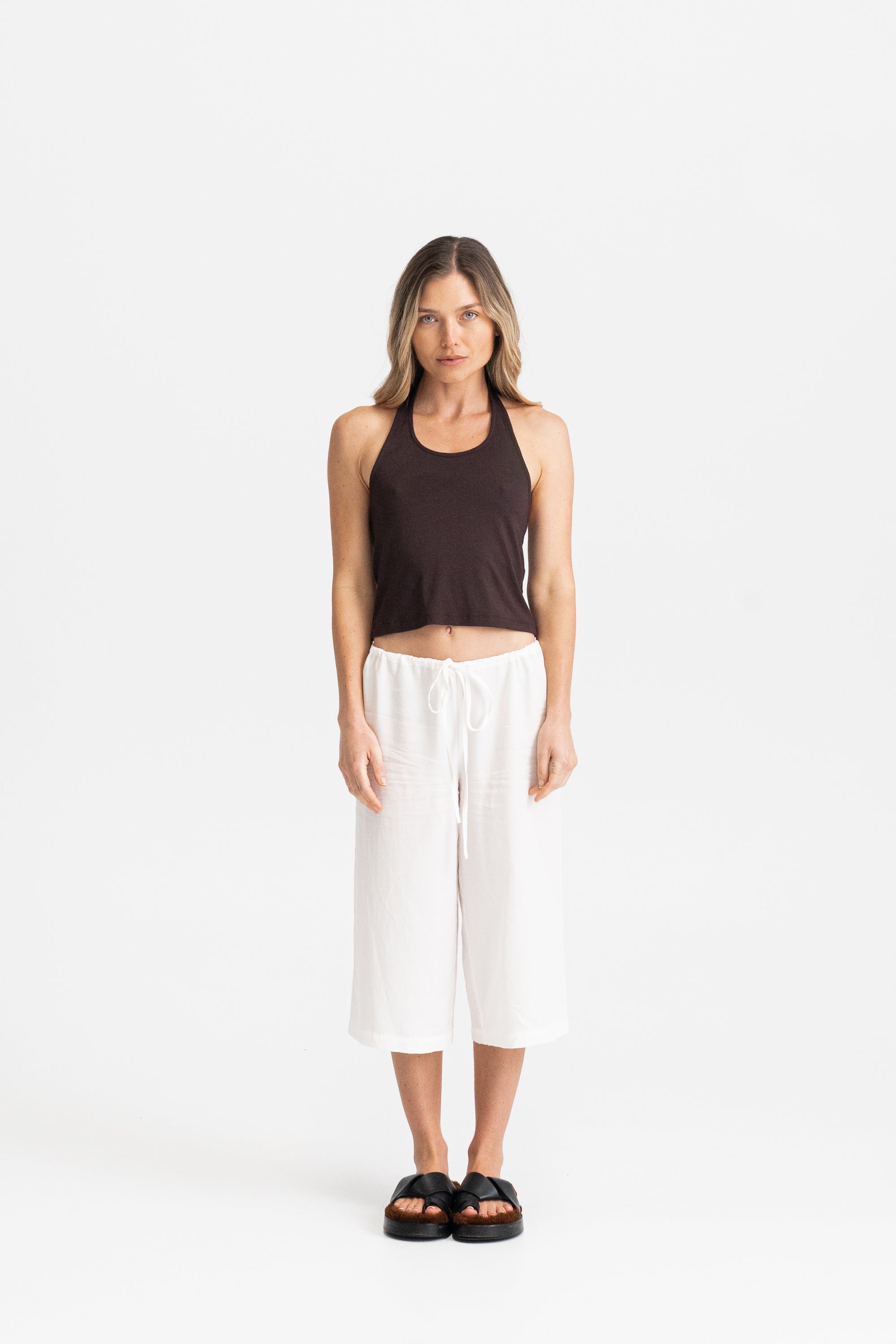 A woman wearing a dark brown modal halter crop top and white wide-leg cropped pants with black slide sandals.