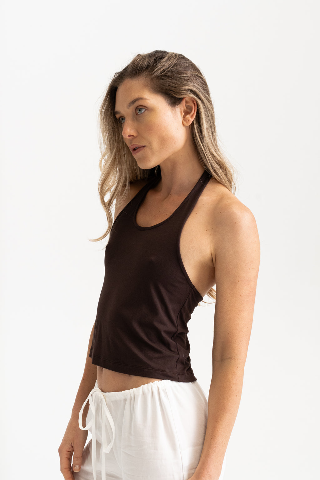 Side view of a woman wearing a dark brown modal halter tank top and white pants.