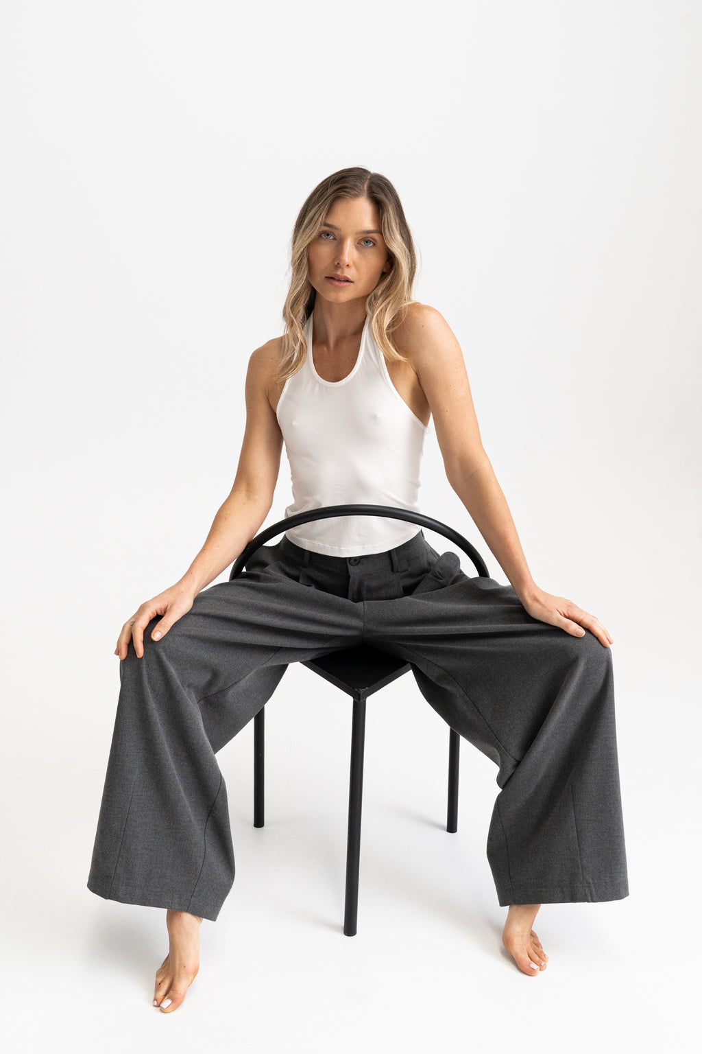 A woman sitting on a black chair, wearing a white sleeveless modal tank top and dark charcoal wide-leg pants.