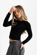 A woman modeling a fitted black long-sleeve cropped button-front top with gray pleated trousers, posed against a clean light gray studio background.