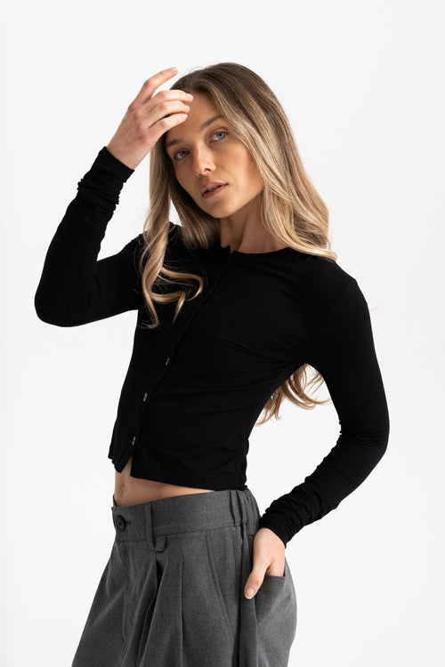 A woman modeling a fitted black long-sleeve cropped button-front top with gray pleated trousers, posed against a clean light gray studio background.