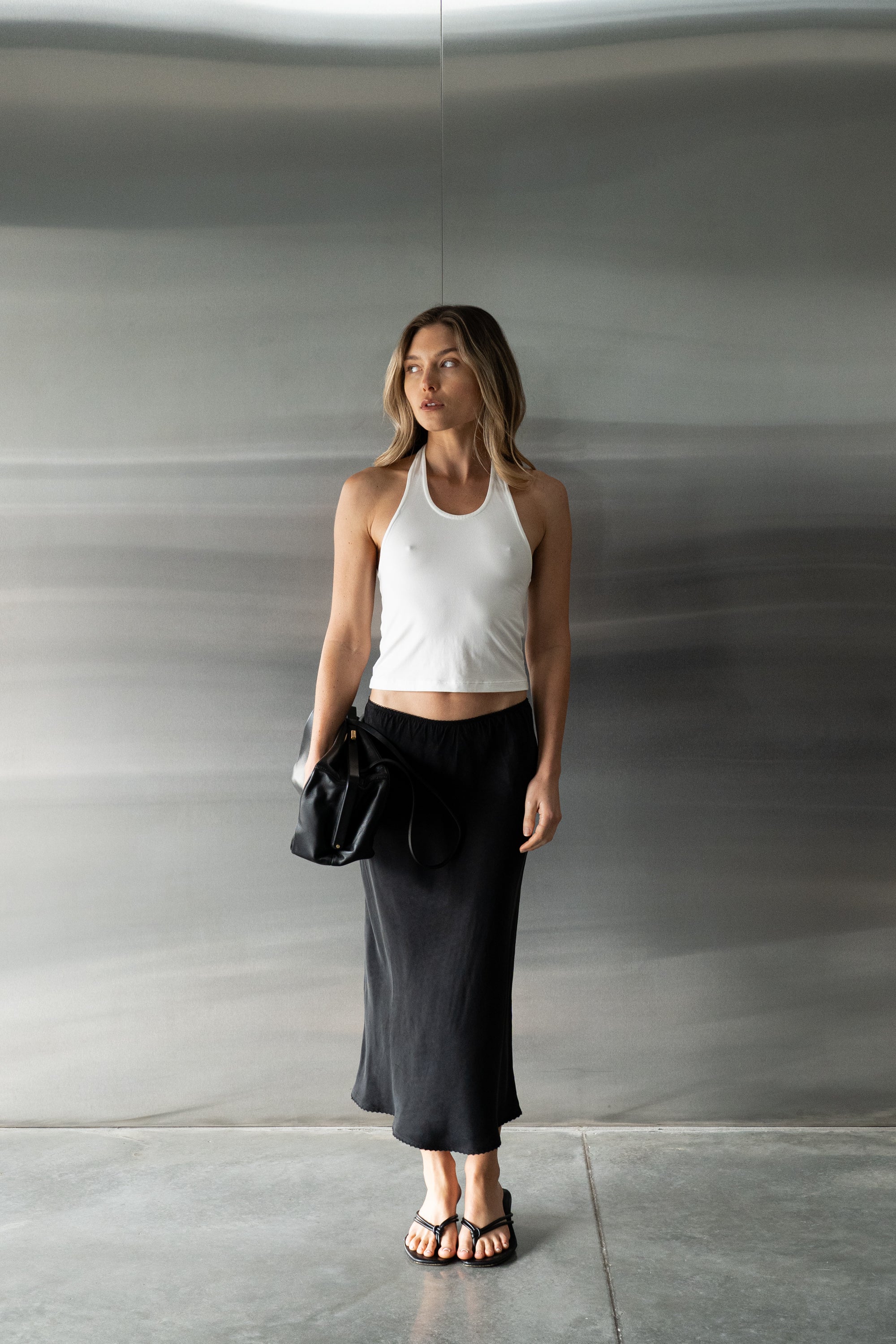 A wearing a white modal tank top, a long black skirt, and black sandals, while holding a black handbag.