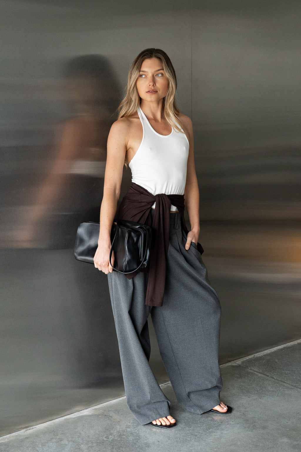 A woman wearing a white modal tank top, loose dark grey wide-leg pants, and a dark brown cardigan wrap tied at her waist. She is holding a black leather clutch.